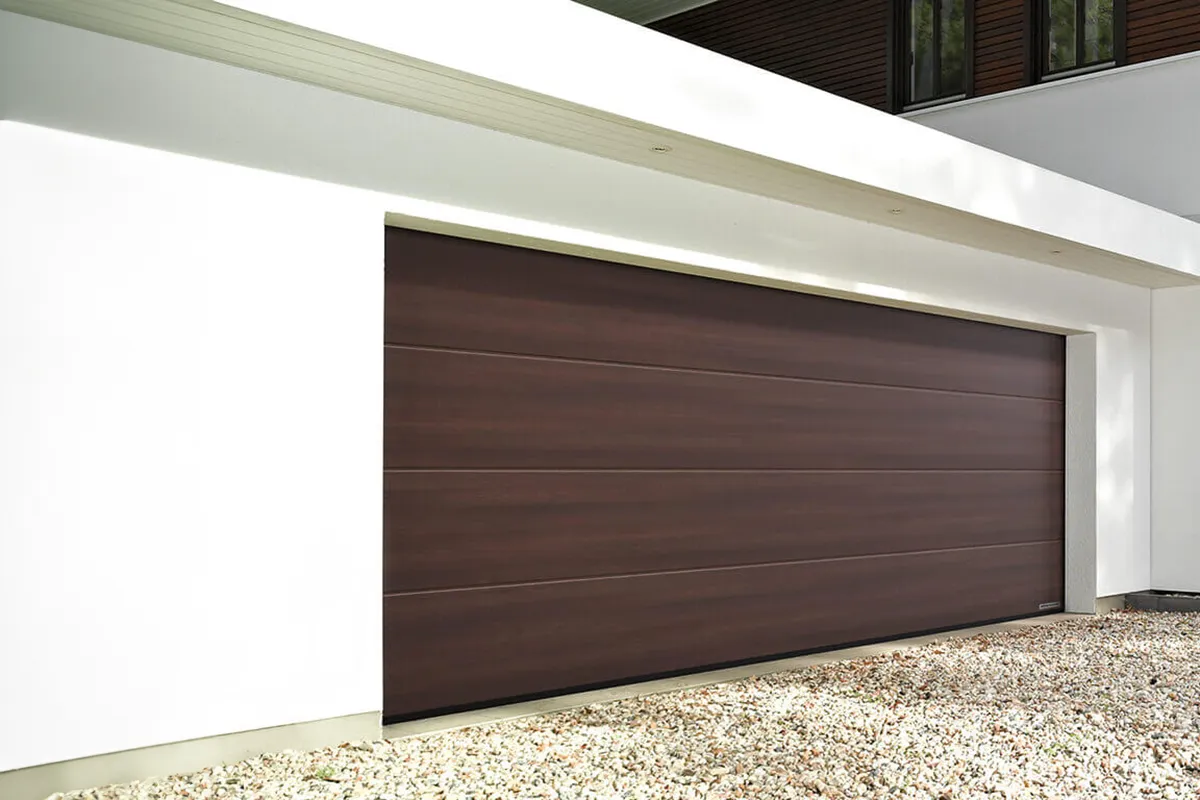 Wysokiej jakości brama garażowa w odcieniu ciemnego drewna, zapewniająca nie tylko bezpieczeństwo, ale i dodająca elegancji zewnętrznym elementom domu, dostępna w ofercie Domo Strefa w Warszawie, gdzie znajdziesz szeroki wybór drzwi zewnętrznych i wewnętrznych.